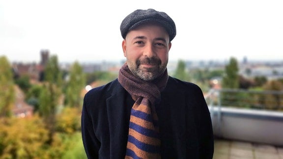Porträtfoto eines Mannes, der eine Mütze und einen Schal trägt. Er steht auf einem Balkon. 