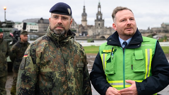 Robert Habermann (l) ,Oberstleutnant der Bundeswehr, und Michael Klahre, Sprecher der Feuerwehr Dresden, stehen am Elbufer nahe der eingestürzten Carolabrücke bei einem Pressegespräch vor der Altstadtkulisse. 