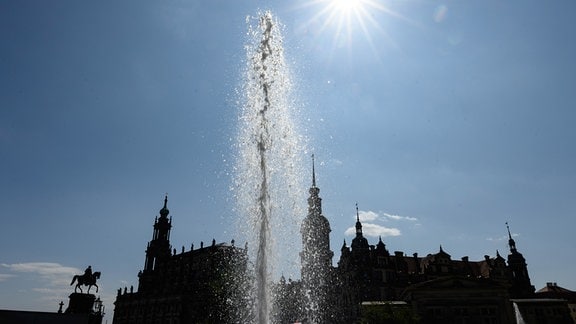 Die Sonne scheint am Himmel über dem Theaterplatz mit dem Reiterstandbild König Johann (l-r), der Hofkirche, dem Hausmannsturm und dem Residenzschloss während die Wasserfontäne eines Springbrunnens aufsteigt.