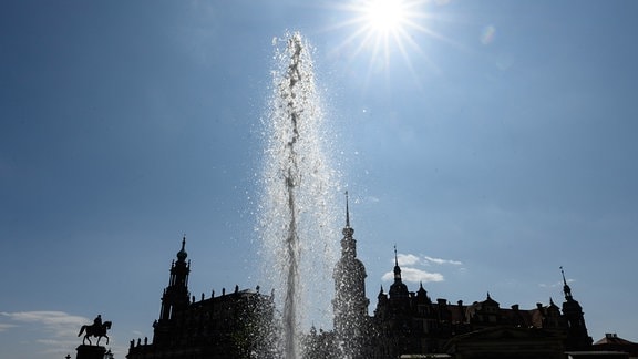 Die Sonne scheint am Himmel über dem Theaterplatz mit dem Reiterstandbild König Johann (l-r), der Hofkirche, dem Hausmannsturm und dem Residenzschloss während die Wasserfontäne eines Springbrunnens aufsteigt.