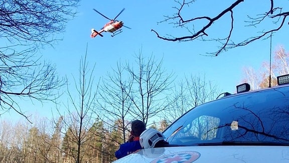 Ein Mann der Bergwacht schaut, wie ein Rettungshubschrauber über Baumwipfeln fliegt.