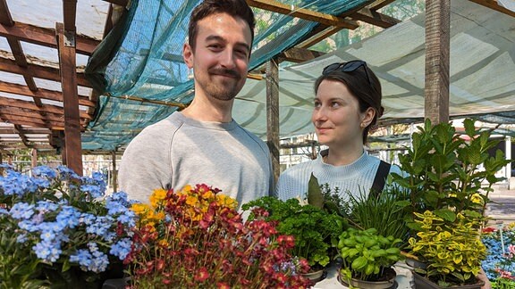 Eine Frau und ein Mann halten Kästen mit Blumen und Kräutern.