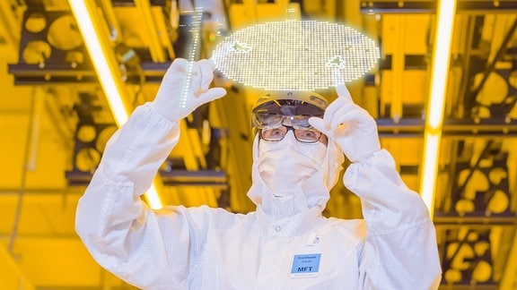 Ein Mann in weißem Overall und Handschuhen steht vor einem transparenten Bildschirm und berührt ihn, der Hintergrund ist gelb.