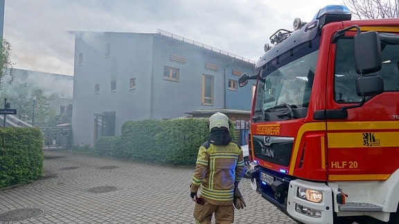 Ein Feuerwehrmann und ein Feuerwehrfahrzeug stehen vor einem Wohnhaus, aus dessen Fenster Rauch steigt.