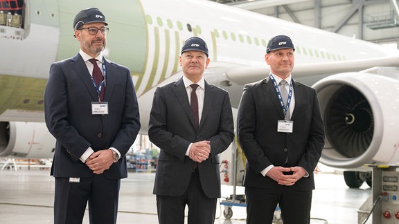 Bundeskanzler Olaf Scholz (SPD, M) steht bei seinem Besuch der Elbe Flugzeugwerke neben Jordi Boto (l), Geschäftsführer Elbe Flugzeugwerke, und Kai Mielenz, Geschäftsführer Elbe Flugzeugwerke, in einen Hangar vor einem Airbus A330.