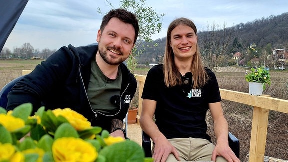 Zwei junge Männer sitzen auf Stühlen an einem Frühlingstag draußen und blicken erwartungsvoll in die Kamera.