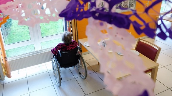 Die Bewohnerin eines Pflegeheimes mit Faschingsdekoration im Vordergrund sitzt in einem Rollstuhl und blickt aus dem Fenster.