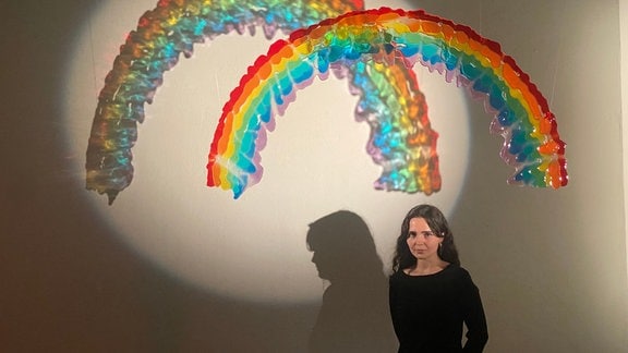 Eine Frau mit langen dunkelbraunen Haaren steht vor einer Kunstinstallation in Regenbogenform. 