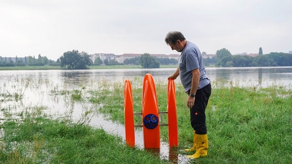 Ein Mann steht mit Gummistiefeln an einem Fluss. Er hält einen kleinen Trimaran, der neben ihm steht. 