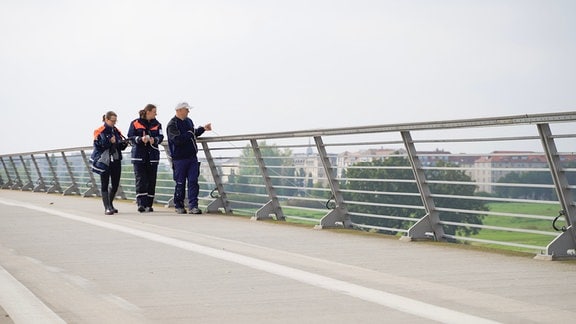 Drei Menschen mit gleicher blau-oranger Arbeitskleidung gehen an einem Brückengeländer entlang. Der Vorderste hält einen Strick, der über das Geländer führt.