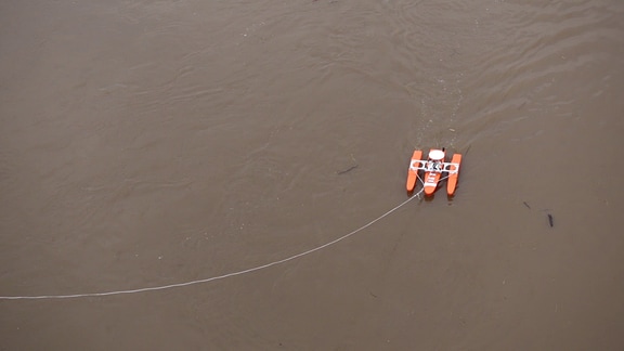 Ein orangefarbenes kleines Boot schwimmt auf braunem Wasser. Es hängt an einem weißen Seil