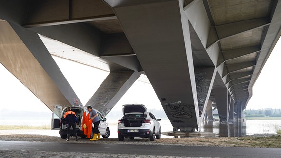 Zwei Autos stehen mit offenen Kofferräumen unter einer Brücke am Fluss. Zwei Menschen räumen ein kleines Plastikboot in einen der Kofferräume.