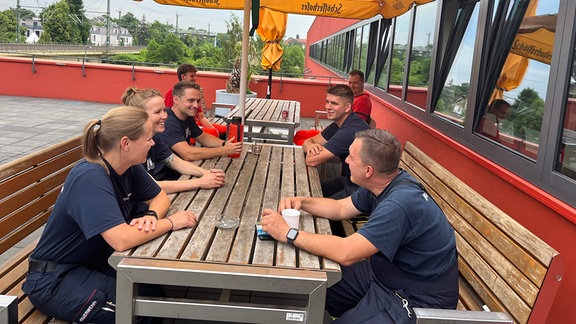 Feuerwehrleute sitzen an einem Tisch unter einem Sonnenschirm.