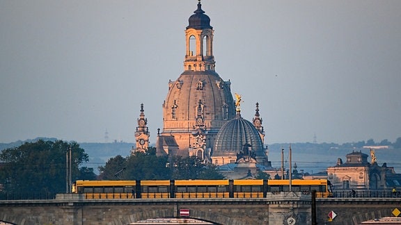 Eine Straßenbahn der Dresdner Verkehrsbetriebe (DVB) fährt am Morgen vor der Altstadtkulisse mit der Frauenkirche (l) und der Kuppel der Kunstakedmie mit dem Engel «Fama» über die Albertbrücke.