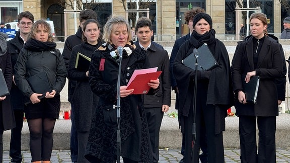 Eine Frau mit weißer Rose am Revers steht vor einem Mikrofon. Hinter ihr stehen Frauen und Männer eines Chores.