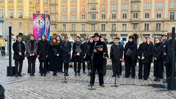 Ein Mann in schwarzer Kleidung steht an einem Mikrofon auf dem Altmarkt in Dresden. Hinter ihm stehen Frauen und Männer eines Chores.