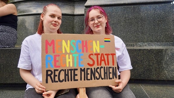 Zwei junge Frauen sitzen am Fuße eines Denkmals und zeigen ein selbstgebasteltes Plakat.