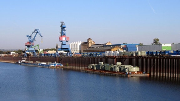 An der hohen Kaimauer des Güterumschlaghafens in Dresden an der Elbe liegt ein Binnenfrachtschiff. An Land stehen unter anderem zwei Verladekräne und zahlreiche Lkw-Sattelauflieger.