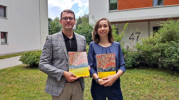 Ein Mann und eine Frau halten ein gelbes Buch in der Hand. Im Hintergrund sind zwei Wohnblöcke zu sehen. 