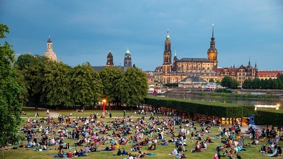 Hunderte Menschen sitzen auf den Wiesen, im Hintergrund die Stadtsilhouette Dresdens mit Frauenkirche, Hofkirche und Schloss.