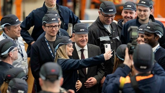 Bundeskanzler Olaf Scholz (SPD m.) macht bei seinem Besuch der Elbe Flugzeugwerke in einem Hangar Selfies mit Mitarbeitern der Flugzeugwerke.
