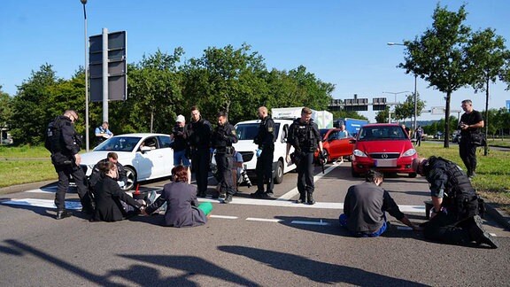 Drei Personen sitzen, von Polizisten umringt, auf einer Straße. Ein Mann hat seine Hand auf der Straße festgeklebt. Im Hintergrund stehen zahlreiche Autos und Transporter.