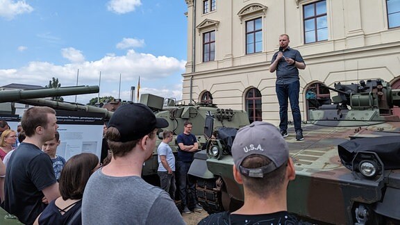 Ein Mann steht auf einem Panzer und spricht zu Museumsgästen. 