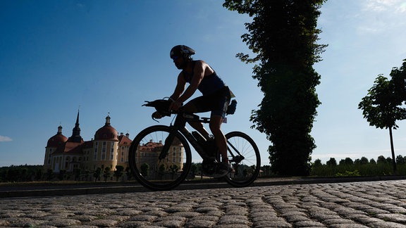 Ein Teilnehmer des Moritzburger Schloss-Triathlon ist vor der Kulisse von Schloss Moritzburg auf seinem Rennrad im Gegenlicht als Silhouette zu sehen. 