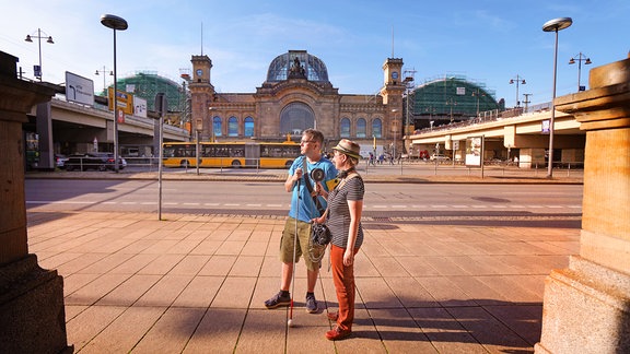 Eine Frau mit Hut und ein Mann mit Taststock stehen vor dem Bahnhofsgebäude in Dresden