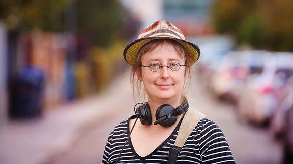Eine Frau mit Brille und Hut steht auf einer Straße. Sie trägt Kopfhörer um den Hals.