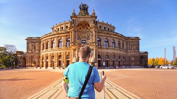 Ein Mann mit türkisem T-Shirt, Taststock für Sehbehinderte und einer gelben Armbinde steht auf dem Platz vor der Semperoper in Dresden.