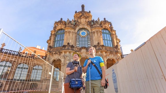 Eine Frau mit Hut hält ein Mikrofon mit Ohren in der Hand, rechts neben ihr steht ein Man mit blauem T-Shirt, Taststock und gelber Armbinde. Im Hintergrund ist ein hohes altes Gebäude zu sehen - der Zwinger in Dresden.