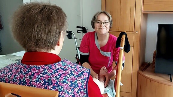 Eine Frau sitzt auf einem Stuhl und hält ihr rechtes Bein einer Altenpflegerin hin. Sie massiert den Unterschenkel vorsichtig.