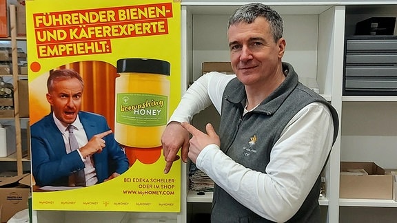 Ein Mann Mitte 40 steht an einem Regal neben einem Plakat. Das Plakat in A3-Größe zeigt den Kopf des Moderators Jan Böhmermann. Der will gerichtlich verbieten lassen, dass der Mann auf dem Foto, ein Imker aus Meißen, mit Böhmermanns Gesichts wirbt.