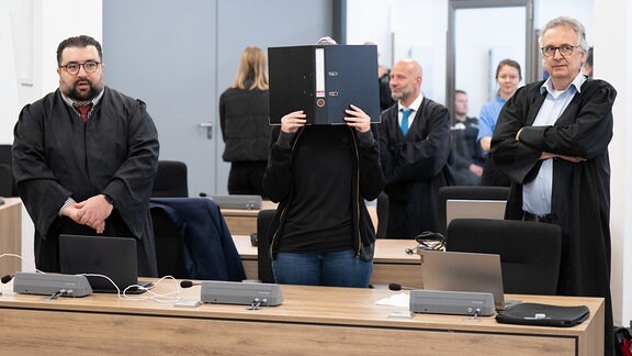 Eine Angeklagte steht bei der Fortsetzung eines Prozesses im Oberlandesgericht Dresden im Verhandlungssaal und hält einen Aktenordner vor ihr Gesicht.
