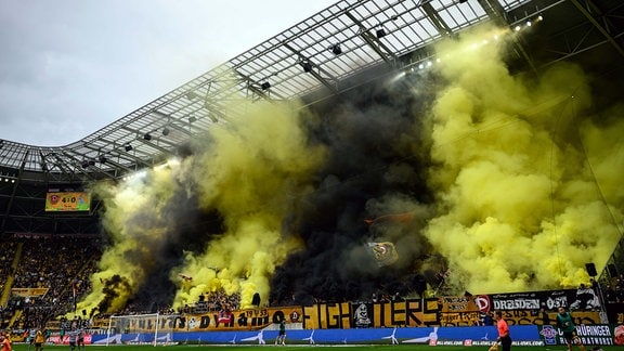 Gelbe und schwarze Rauchwolken über Fußballfans im Stadion.