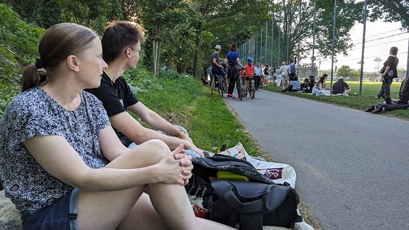Ein Mann und eine Frau sitzen am Rand eines Weges an der Wiese. Im Hintergrund stehen und sitzen weitere Menschen an einem Zaun.