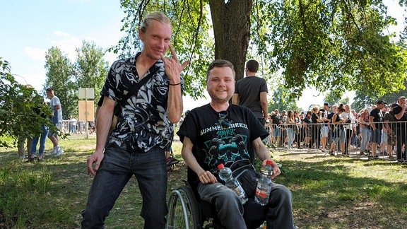 Ein Mann im Rollstuhl und ein anderer Mann lachen in die Kamera.