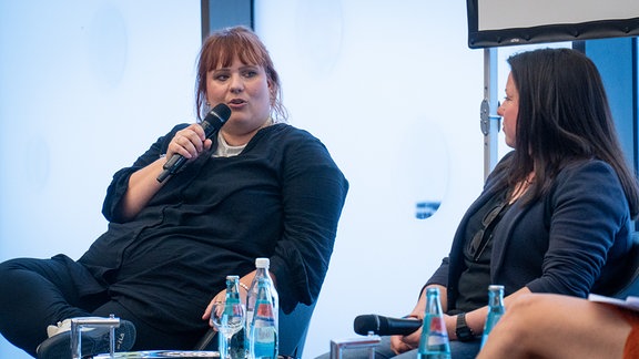 Eine Frau mir rötlichen, zurückgebundenen Haaren sitzt auf einem Podium und spricht in ein Mikrofon.