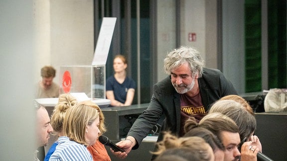 Ein Mann mit grauen Locken und grauem Bart und Anzug hält einer jungen, blonden Frau ein Mikrofon hin.