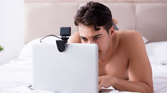 Junger Mann liegt mit nacktem Oberkörper auf einem Bett und schaut auf einen Laptop mit Kamera.