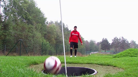 Ein Fußball springt in ein überdinensionales Golf-Loch mit Fahne, dahinter beobachtet ein Fußballer seinen Schuss.