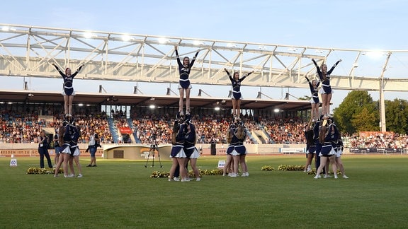 Eine Gruppe Cheerleader macht vier Hebefiguren auf einer Wiese vor einer gefüllten Publikumstribühne.