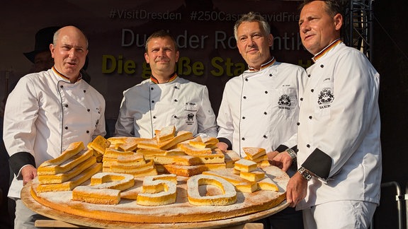 Auf dem Neumarkt Dresden stehen vier Bäckermeister mit einen großen Holzbrett. Darauf liegt Kuchen in Form einer 250 zum 250. Geburtstag von Caspar David Friedrich.