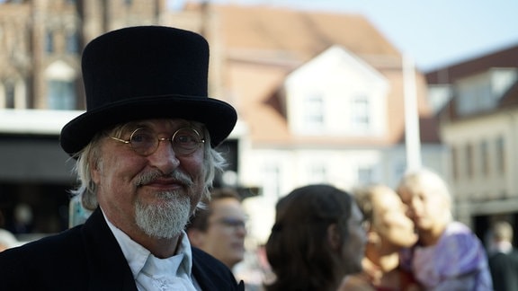 Auf dem Markt in Greifswald in Mecklenburg-Vorpommern steht ein älterer Mann mit einem stattlichen Zylinder.