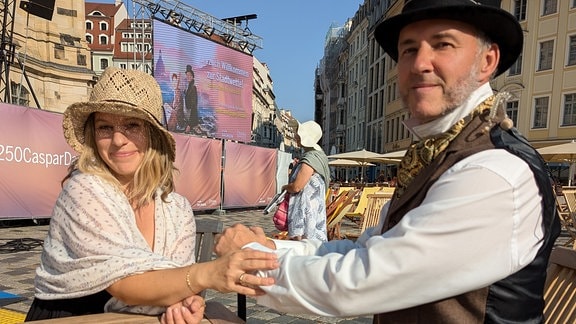 Die beiden MDR SACHSEN-Moderatoren Silvio Zschage (rechts) und Elena Pelzer haben sich in romantische Kleider begeben und warten an einem Tisch auf dem Neumarkt Dresden auf das Ergebnis der Wette.