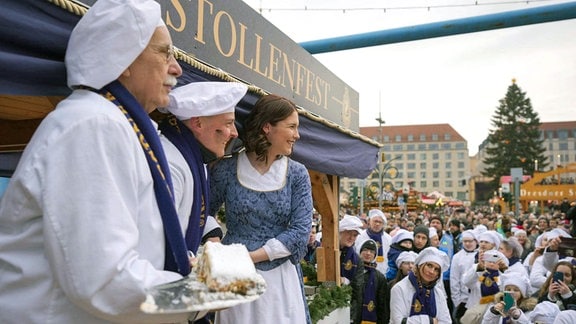 Eine junge Frau im Kleid und zwei Männer mit Bäckermützen stehen auf einer Bühne. Einer der Männer trägt ein Stück Stollen auf einem Tablett. Im Hintergrund sind zahlreiche Menschen zu sehen.