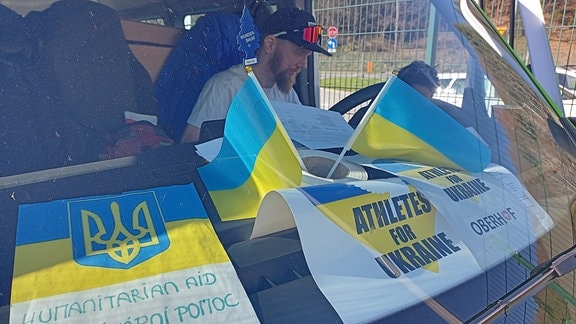 Mann mit Basecap und Sonnenbrille sitzt in Transporter, dessen Armaturenbrett ist mit ukrainischen Flaggen übersät