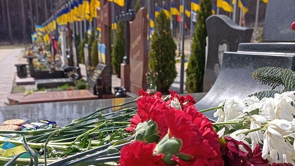 Blumen auf Grabsteinen, hinter den ukrainische Flaggen wehen - in Irpin