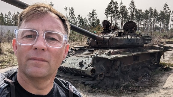 Mann mit Brille blickt nachdenklich und steht vor Wrack eines Panzers